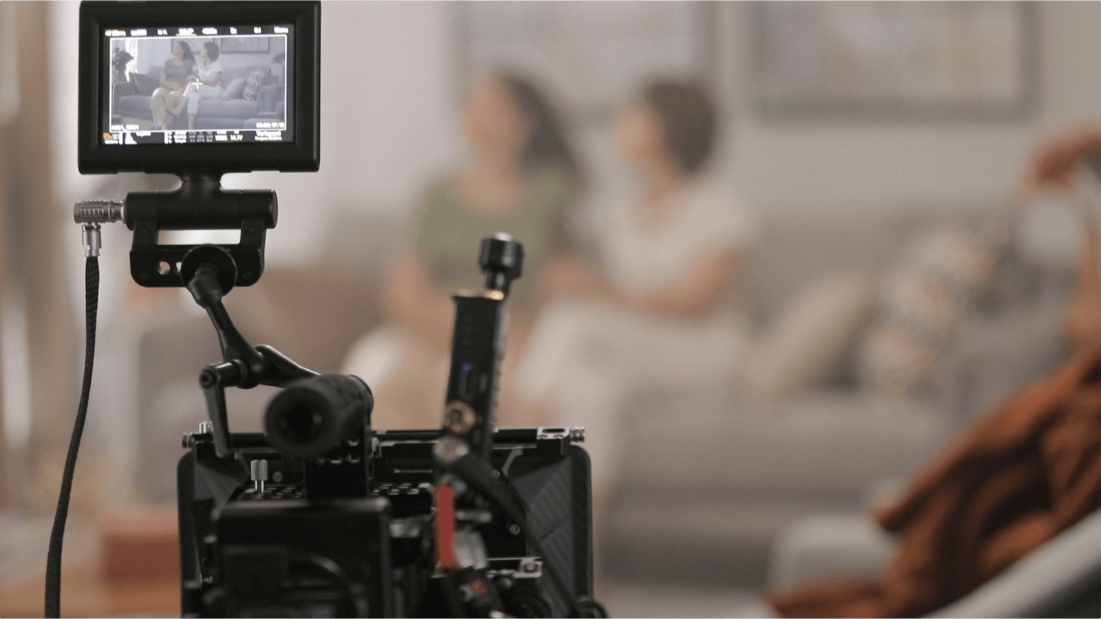 Câmera de filmagem cinematográfica de tamanho médio, filmando uma cena que acontece ao fundo com duas pessoas sentadas em um sofá em uma sala de casa. Essa cena é parte de uma produção da Cria Filmes, uma produtora audiovisual de Curitiba.